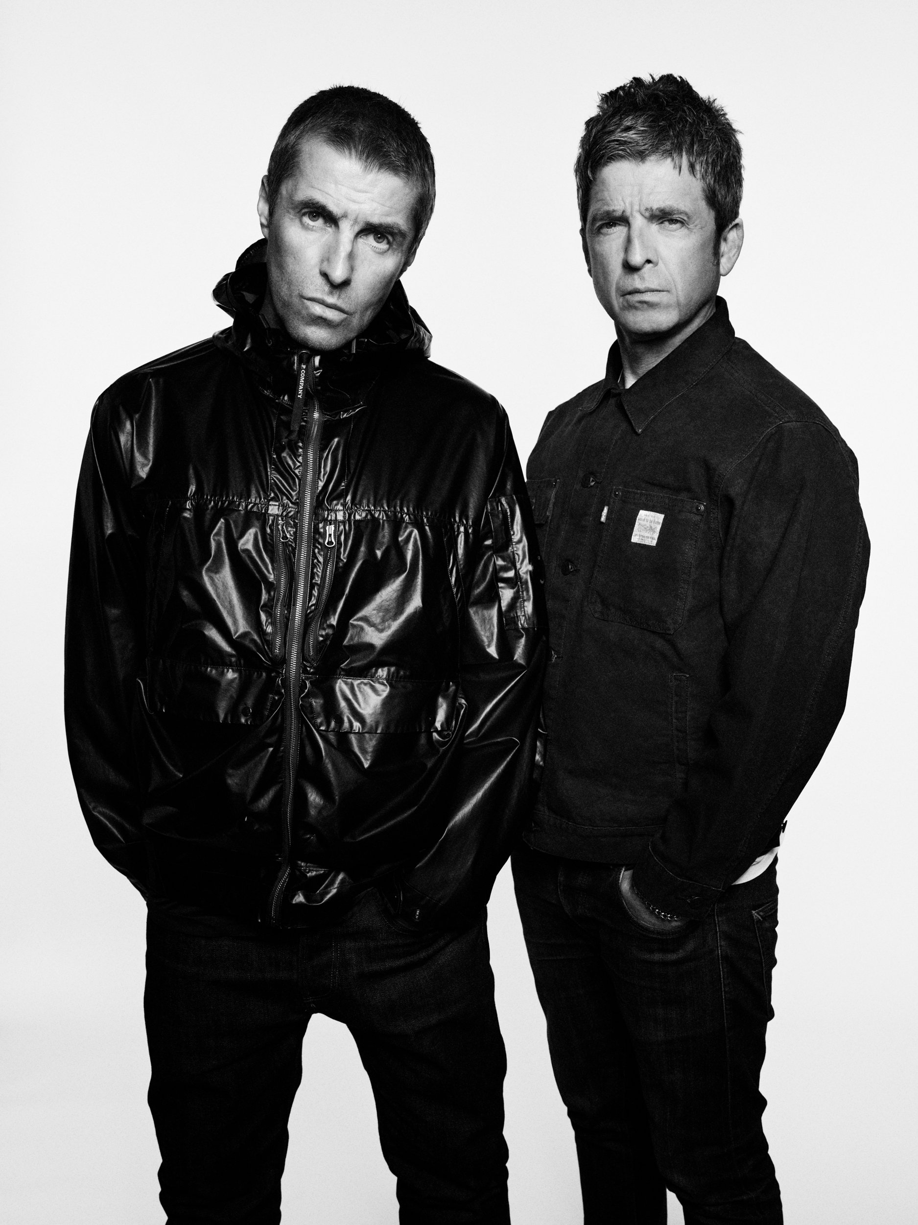 Liam and Noel Gallagher pose in all black outfits, in a black and white Oasis promo image